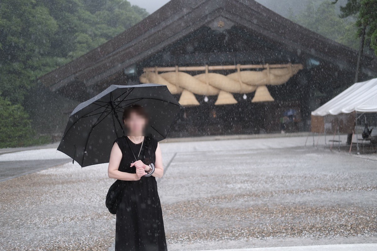 激しい雨の中、傘をさして神楽殿の前に立つ参拝者。奥に神楽殿の大注連縄が見える